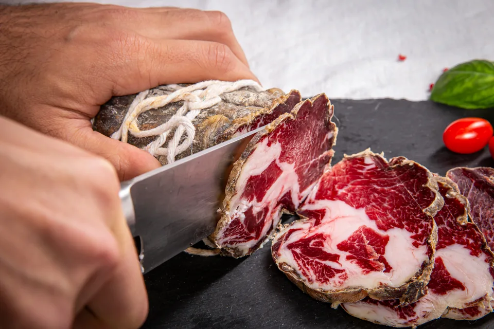 Des mains tranchent une coppa au couteau sur une ardoise.