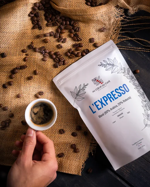 Un sachet de café « L'Expresso » et une tasse d'expresso tenue en main, sur une toile de jute.