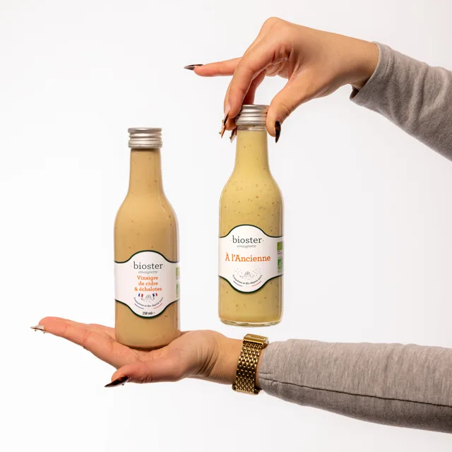 Deux bouteilles de vinaigrettes Bioster tenues en main sur fond blanc.