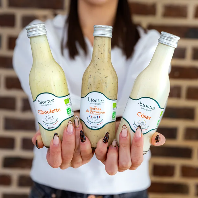 Trois bouteilles de sauces Bioster présentées à deux mains devant un mur de briques.