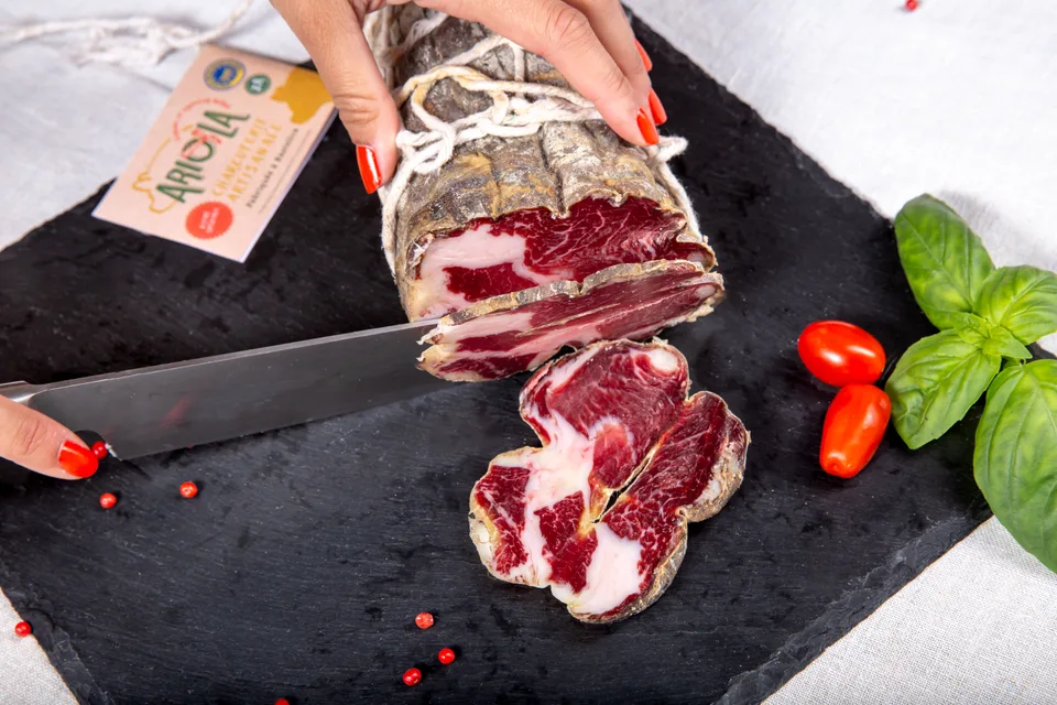Une coppa tranchée au couteau sur une ardoise, avec basilic et tomates cerises.