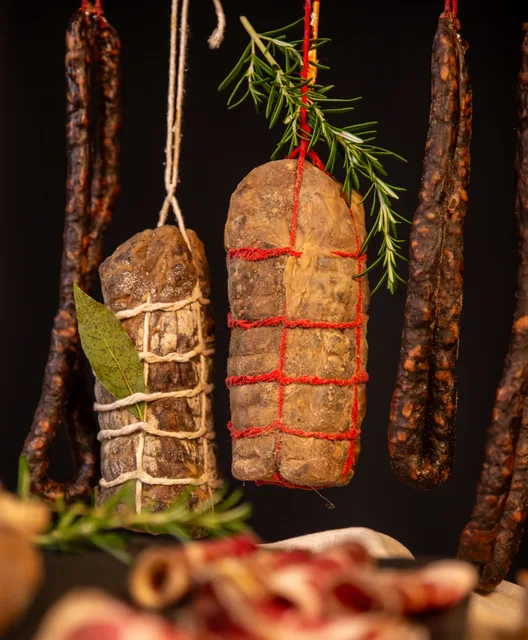 Des saucissons et une pièce de charcuterie suspendus, sur fond sombre.